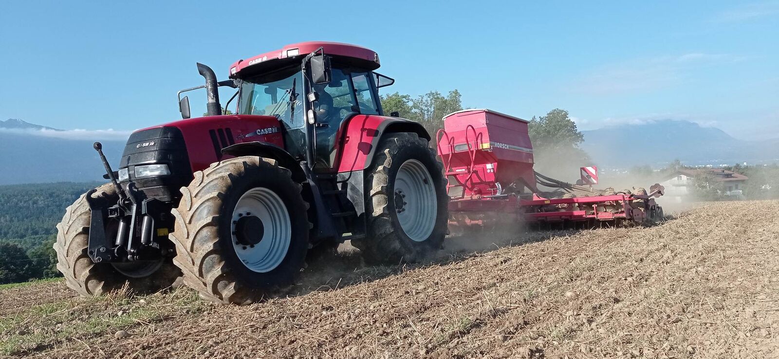 Case IH Traktor mit Horsch Sämaschine bei der Aussaat im Morgenlicht, Staubfahne vor Kärntner Bergpanorama